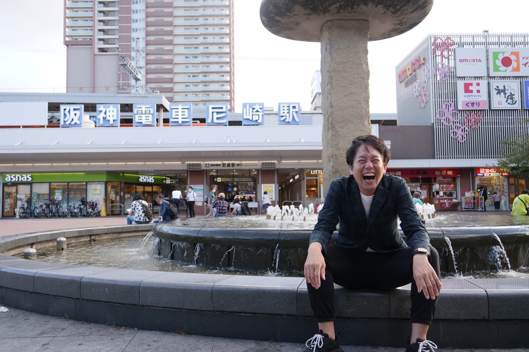 写真、阪神尼崎駅北側の噴水の縁に座る笑顔の成海さん