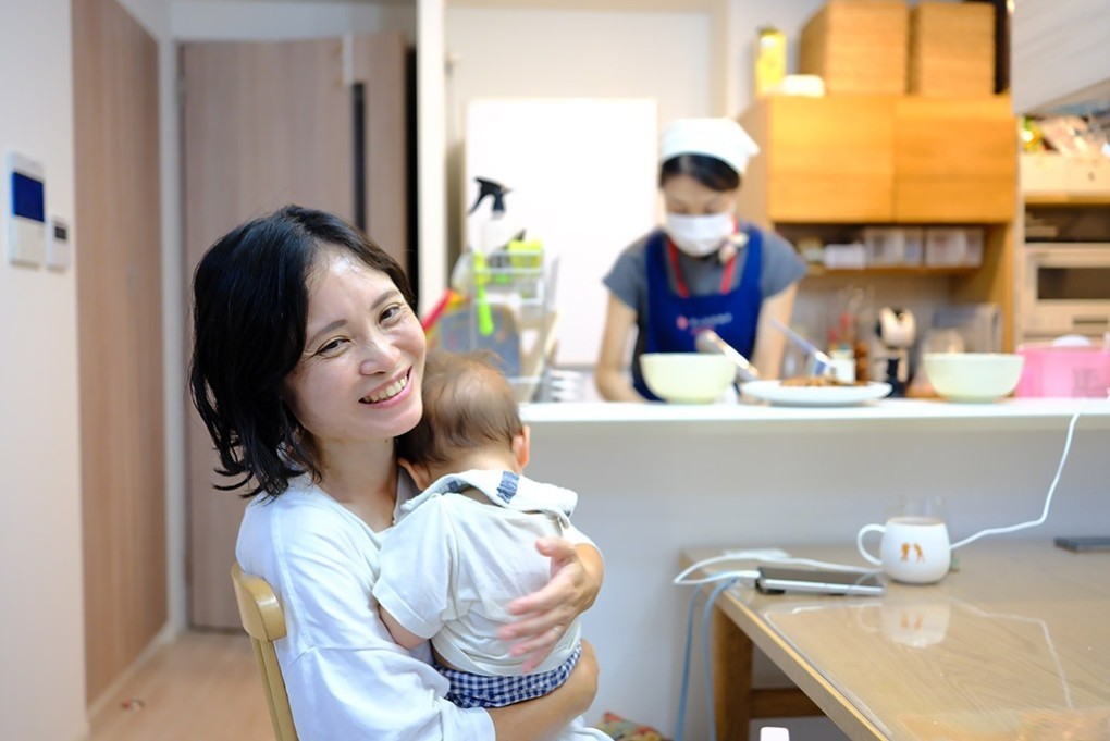 写真、赤ちゃんを抱いて話す利用者。奥のキッチンでヘルパーが料理を作る