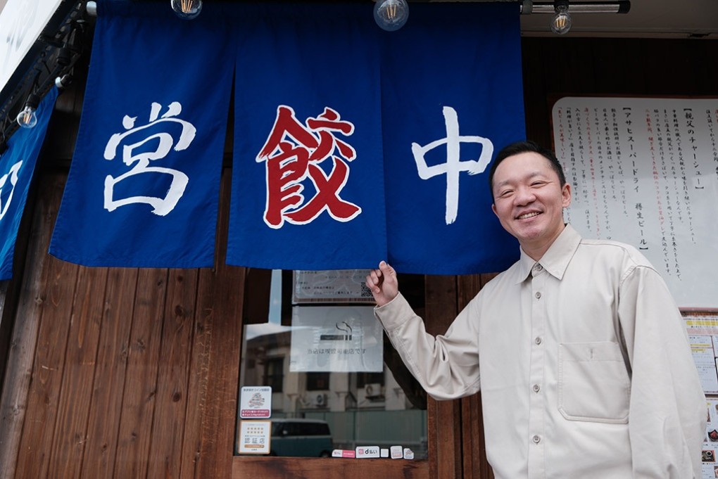 写真、のれんを手に笑顔の辻さん