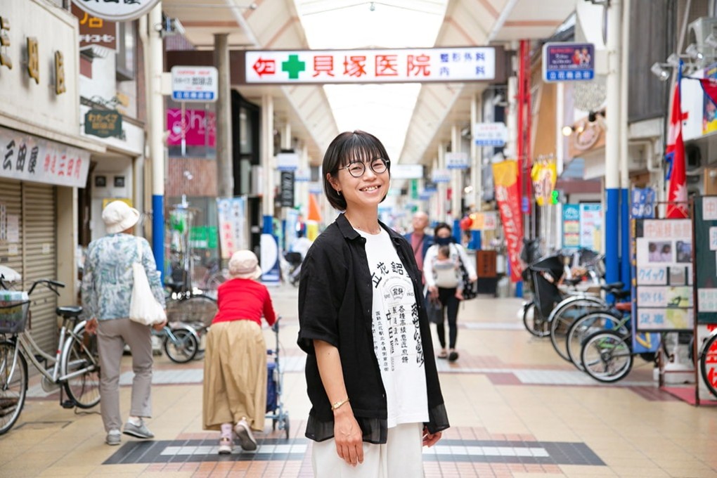 写真、立花商店街の中で笑顔の黒田さん。中に中田屋さんのオリジナルTシャツを着込んでいる。